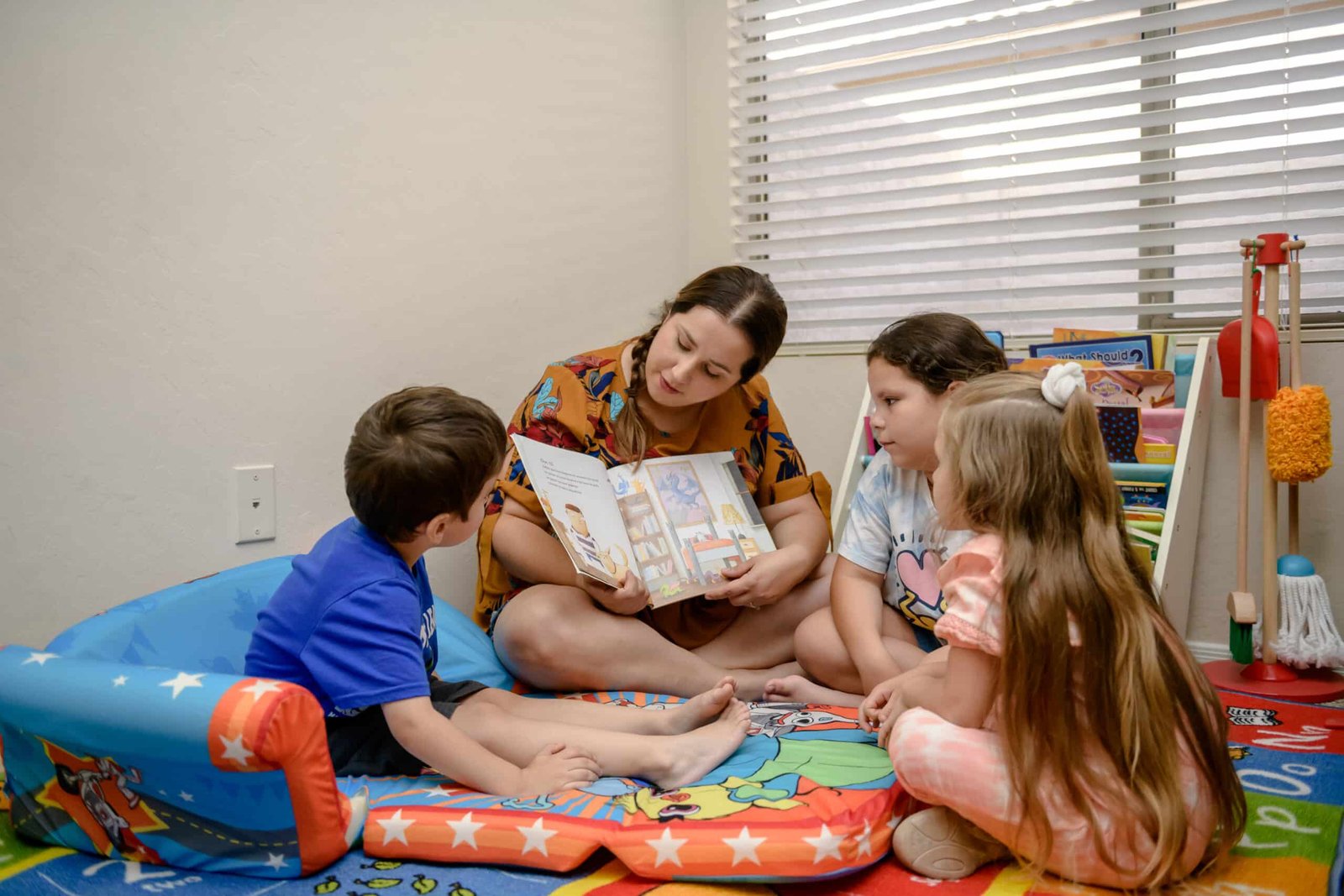 A home daycare provider reading a picture book to three children in a cozy reading corner